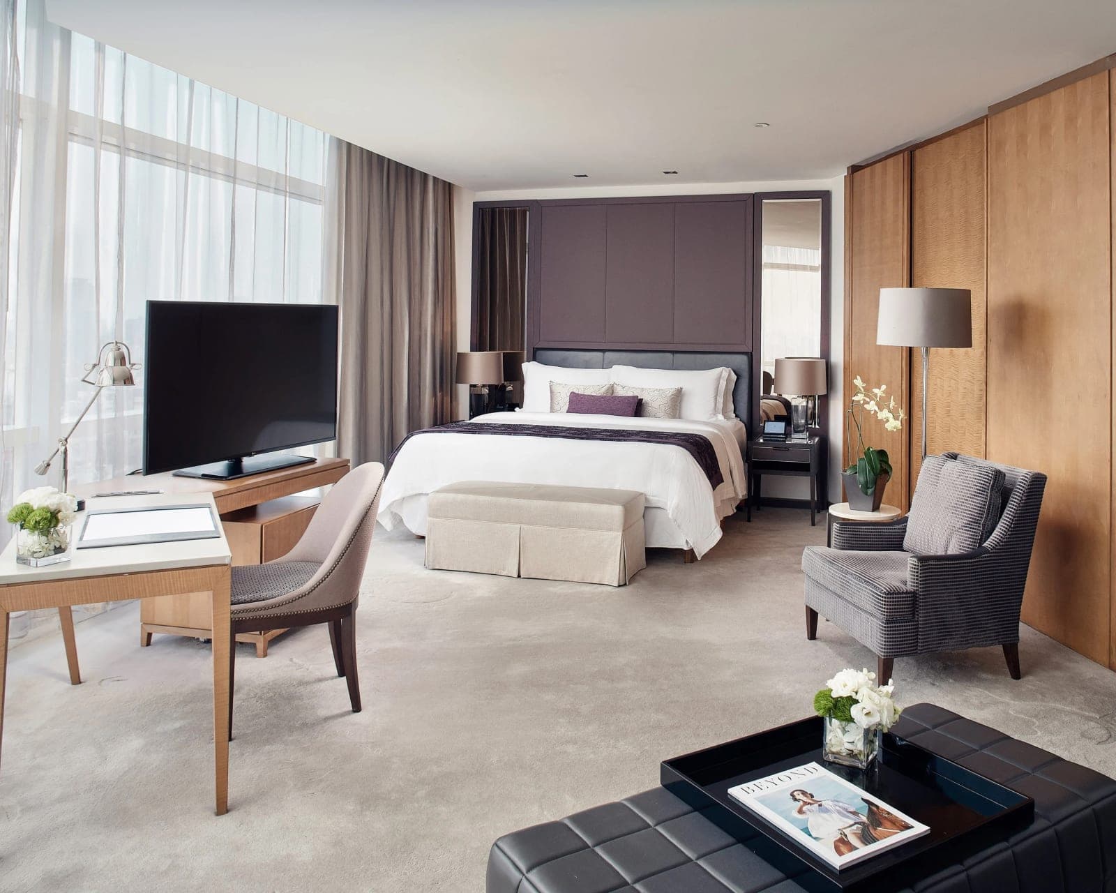 a hotel suite with wood and brown walls, windows, white carpet, bed at the far side with chairs and desk in the foreground