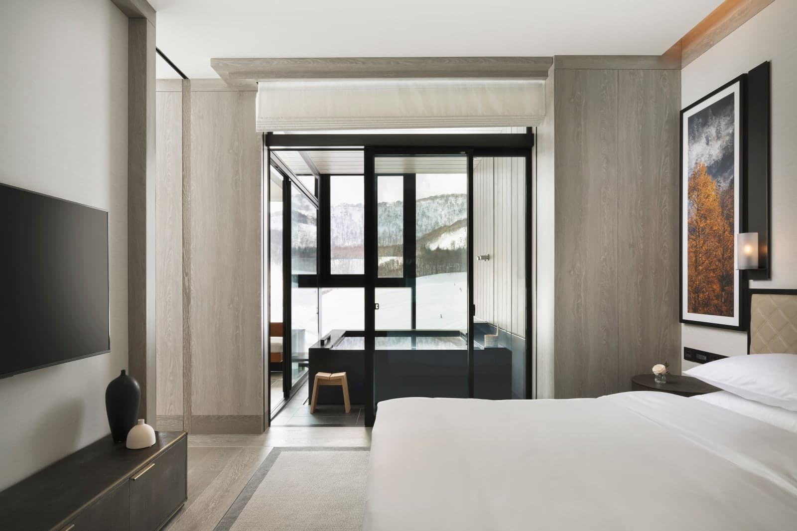 Sleek, minimalist hotel bedroom with a king bed, warm wood paneling, and a glass-enclosed soaking tub overlooking snow-covered mountains.
