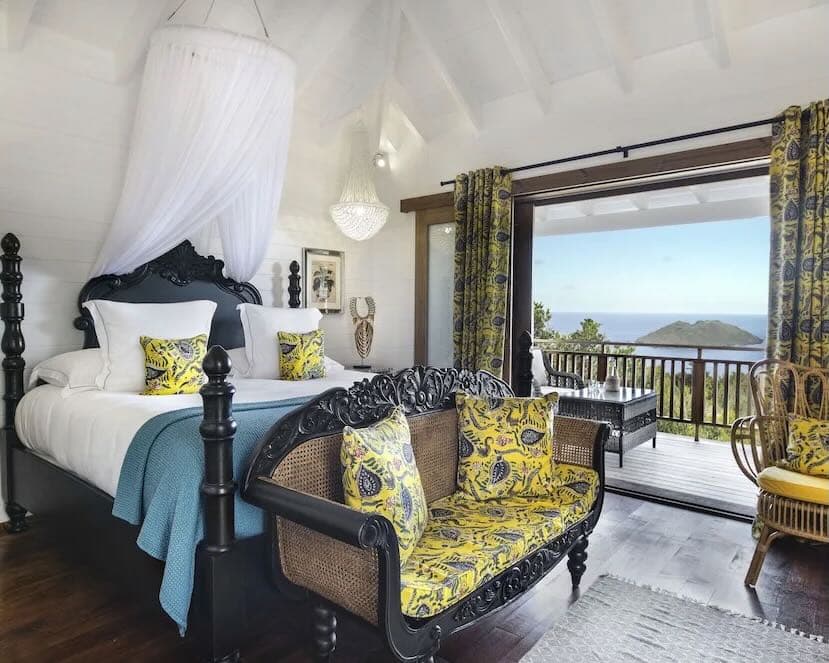 an eclectic hotel room with black four poster bed, teal banket, yellow and green accent pillows, chairs, and drapers overlooking the ocean