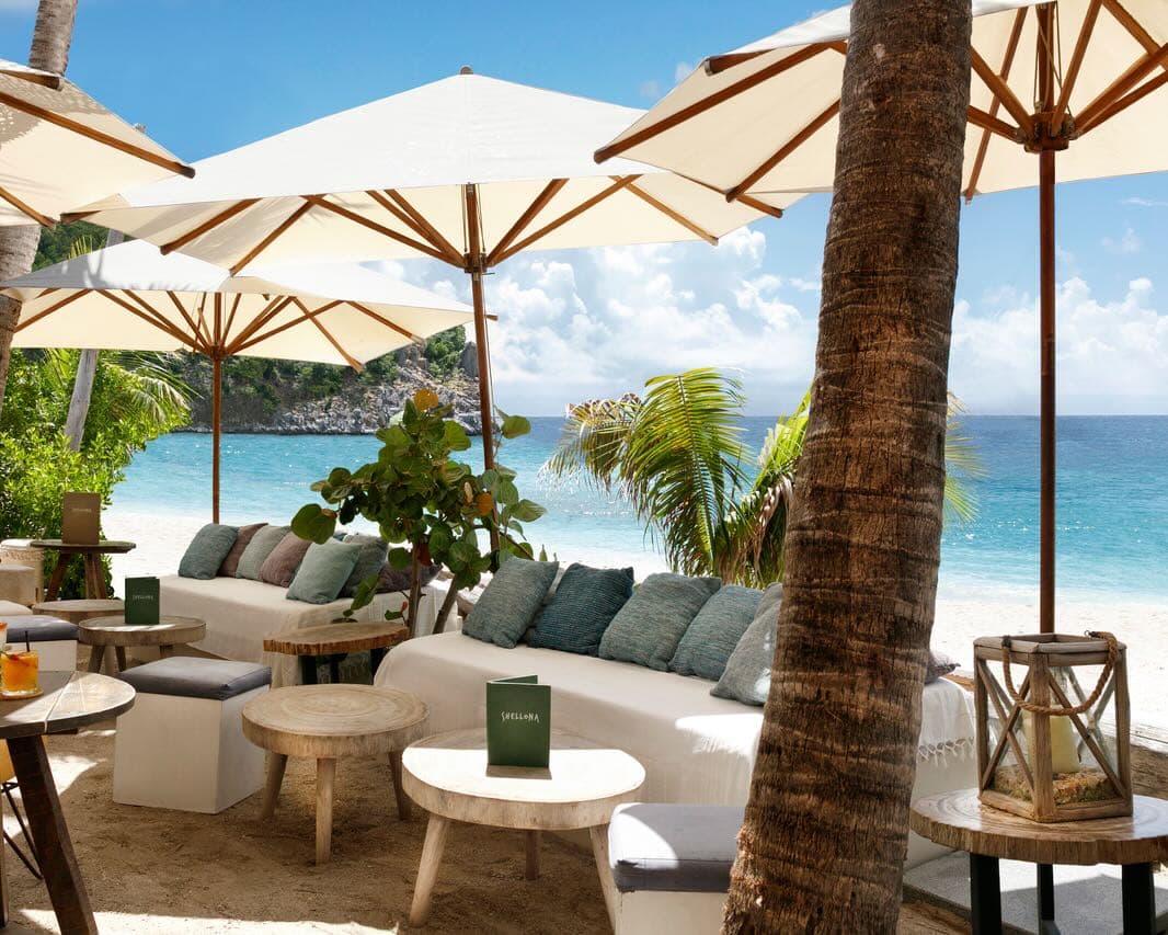 a beachside lounge with sofas, side tables, and umbrellas next to white sand and turquoise waters during day