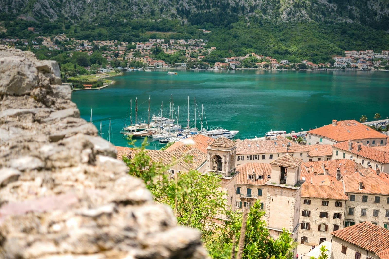 Aerial view of a coastal town with terracotta-roofed buildings, a marina with sailboats, and green hills in the background.