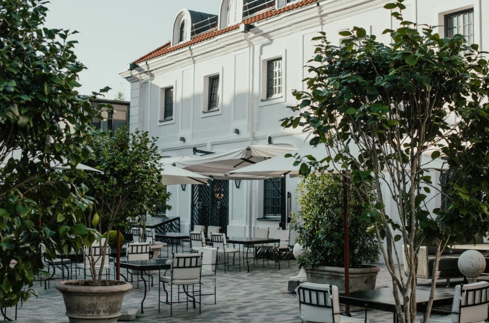 Outdoor courtyard with black tables and white cushioned chairs, large potted plants, umbrellas, and a white building in the background.